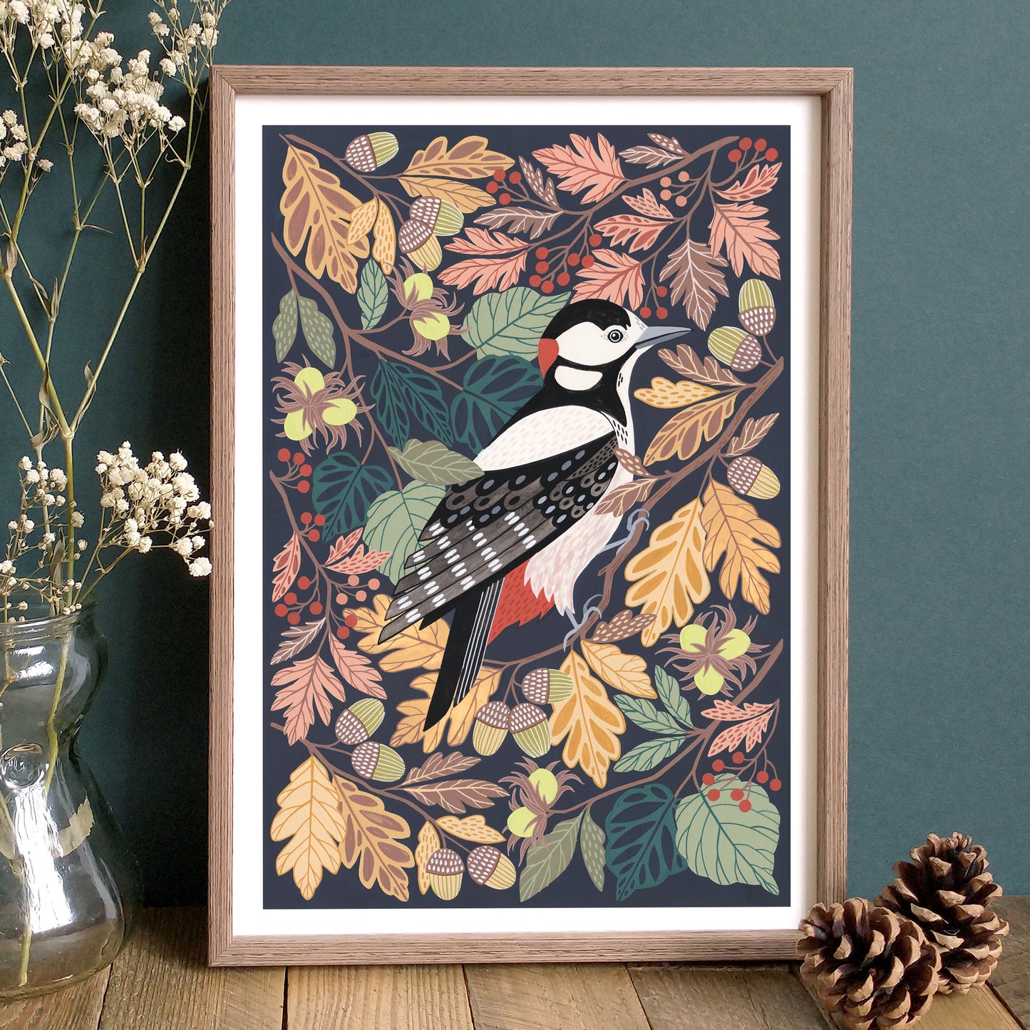 Framed artwork of a woodpecker among autumn leaves and berries on a wooden surface.