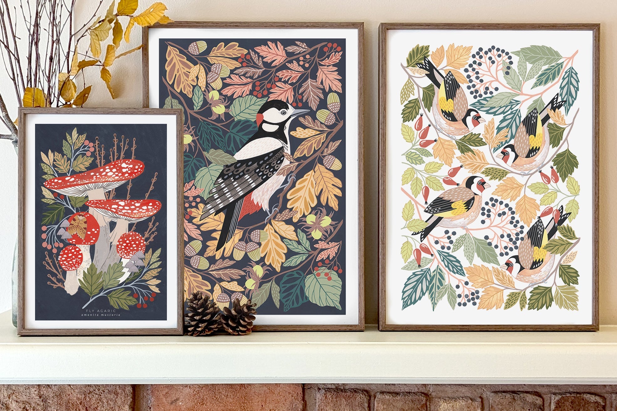 Three framed woodland art prints displayed above a brick fireplace. The prints feature a woodpecker, red-and-white mushrooms, and birds among autumn leaves.