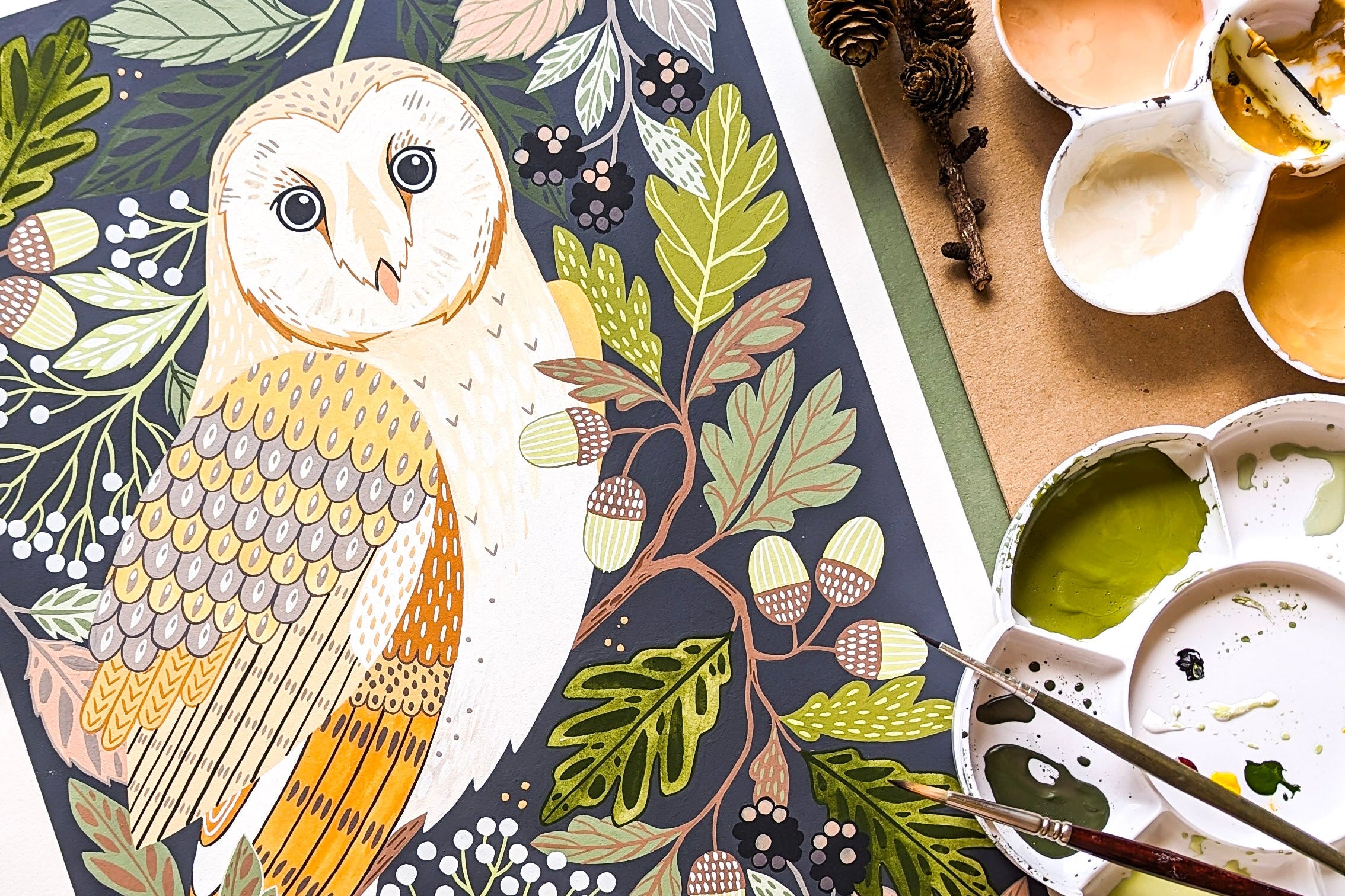 A detailed illustrated painting of a barn owl surrounded by autumn leaves, acorns, and berries, shown in a styled flat-lay with paint palettes, brushes, and pinecones.