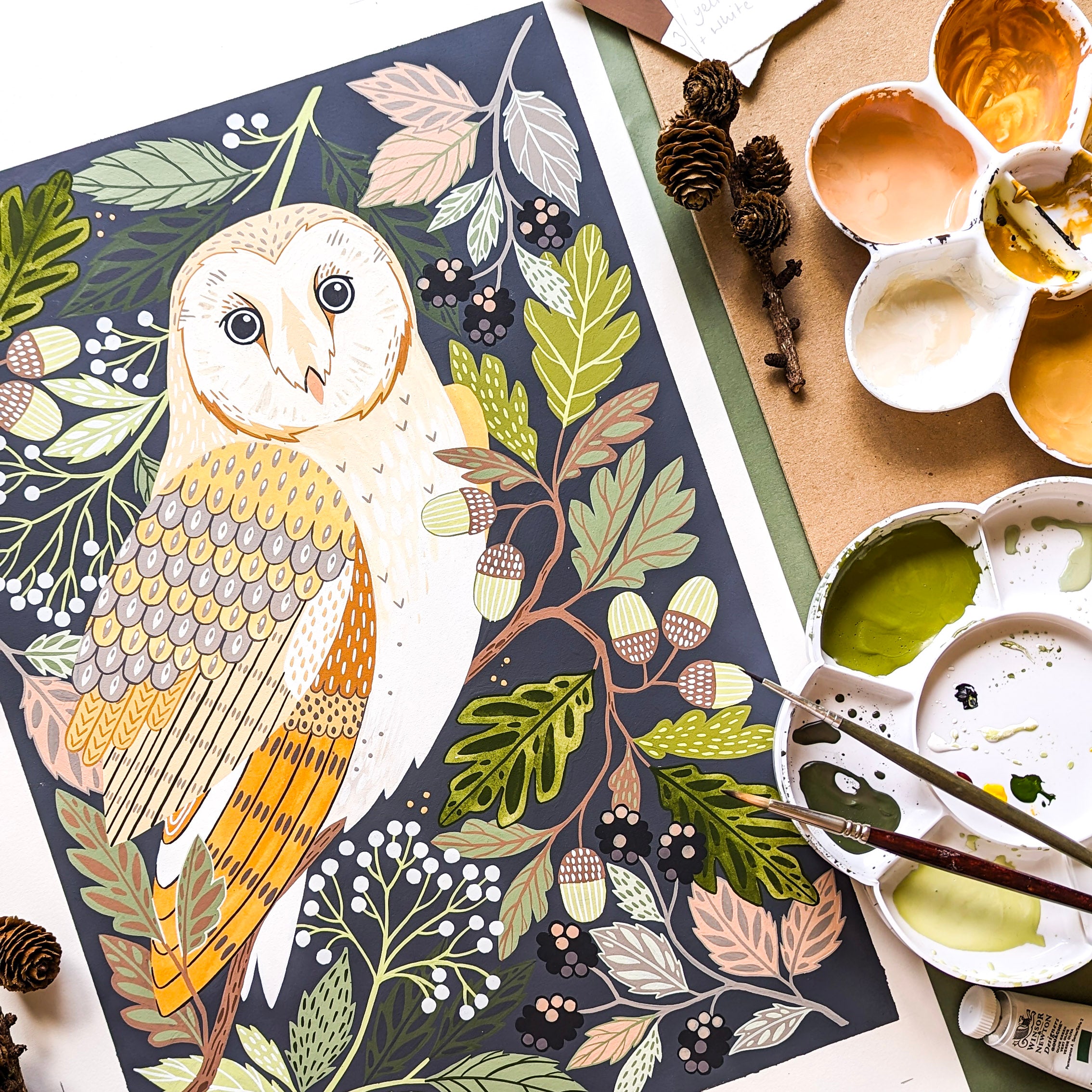 A detailed illustrated painting of a barn owl surrounded by autumn leaves, acorns, and berries, shown in a styled flat-lay with paint palettes, brushes, and pinecones.