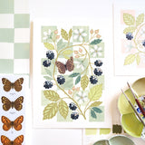 A botanical art print featuring blackberries, white flowers, and a butterfly on a chequered background, styled in a flat-lay with paintbrushes, a paint palette, and other art prints.