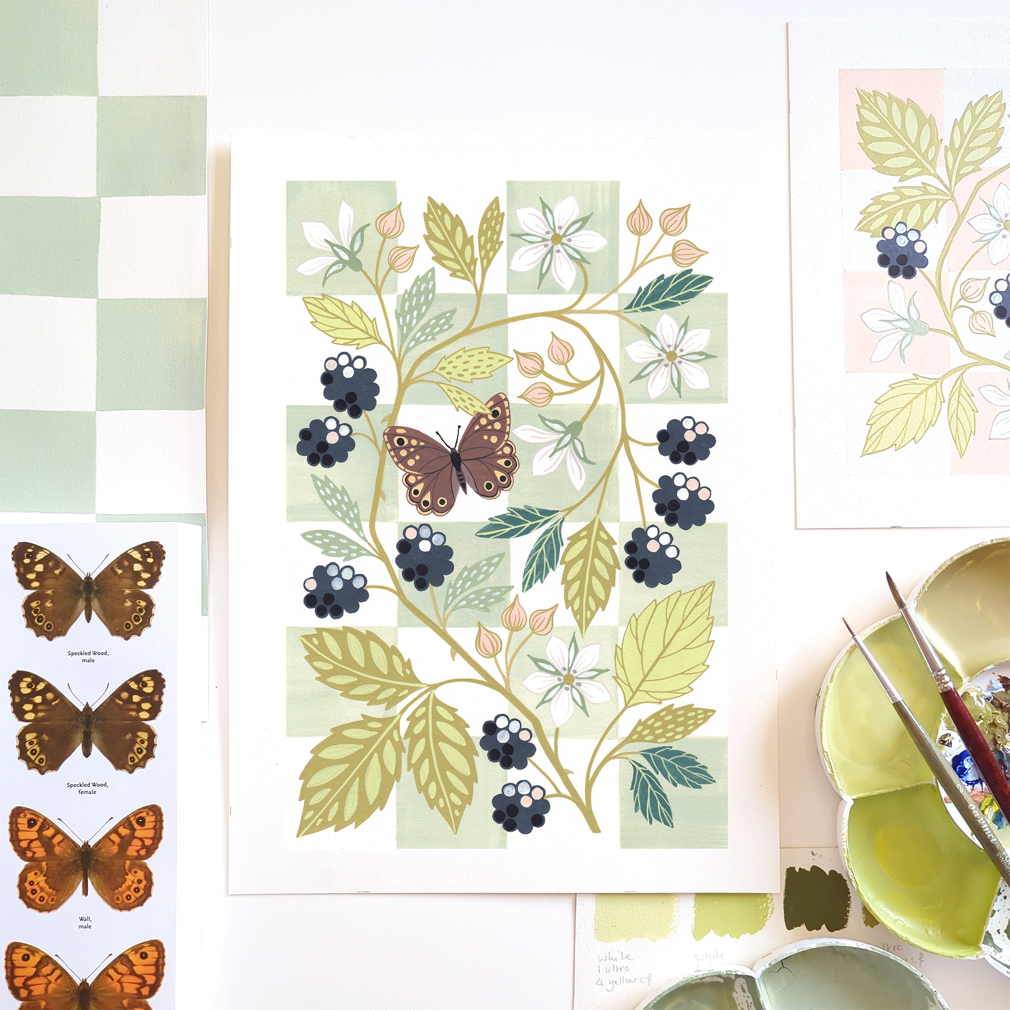 A botanical art print featuring blackberries, white flowers, and a butterfly on a chequered background, styled in a flat-lay with paintbrushes, a paint palette, and other art prints.