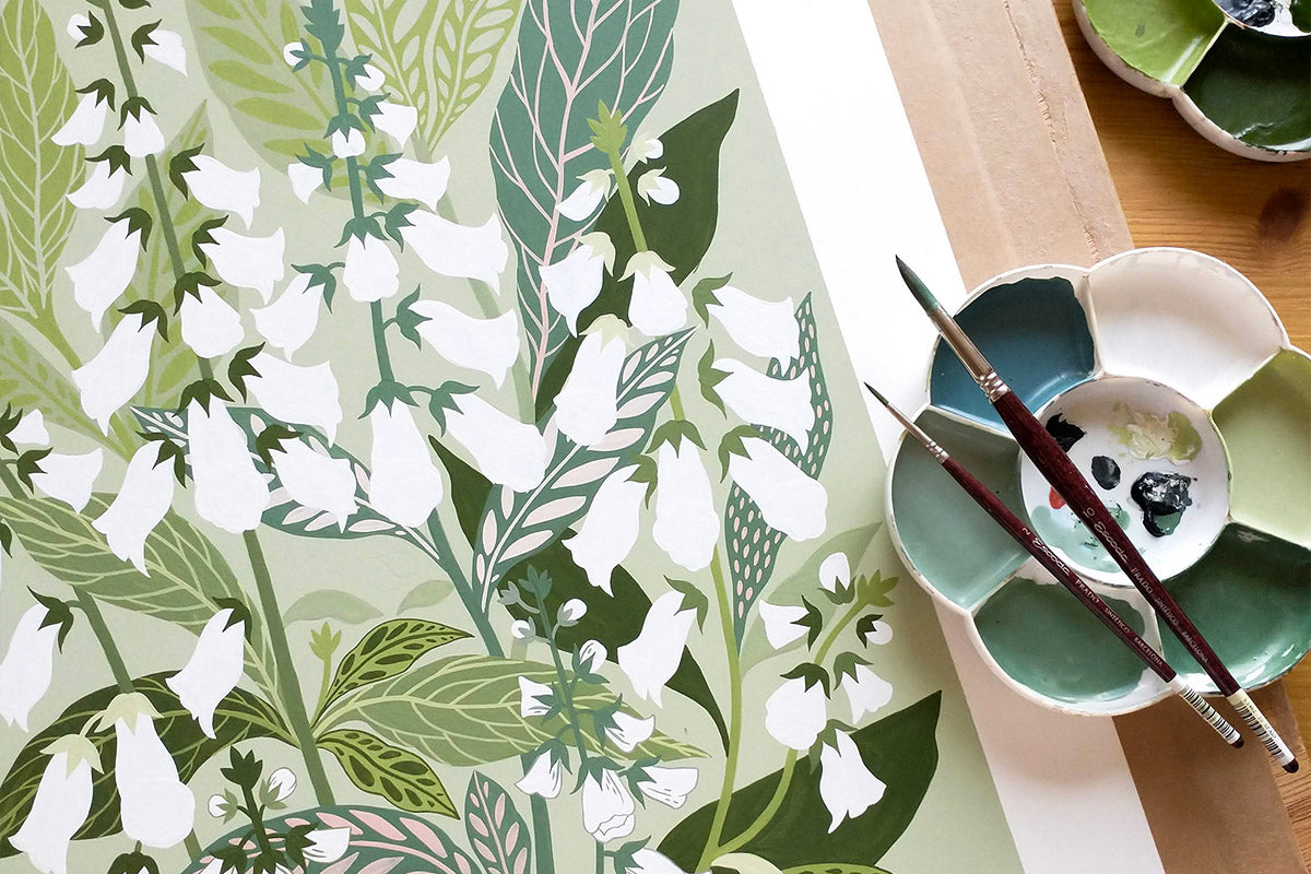 A close-up of a botanical painting featuring white foxglove flowers and green leaves on a pale sage background, styled with a paint palette and paintbrushes on a wooden surface.