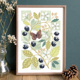 A framed botanical art print featuring a butterfly, leaves, flowers, and blackberries.