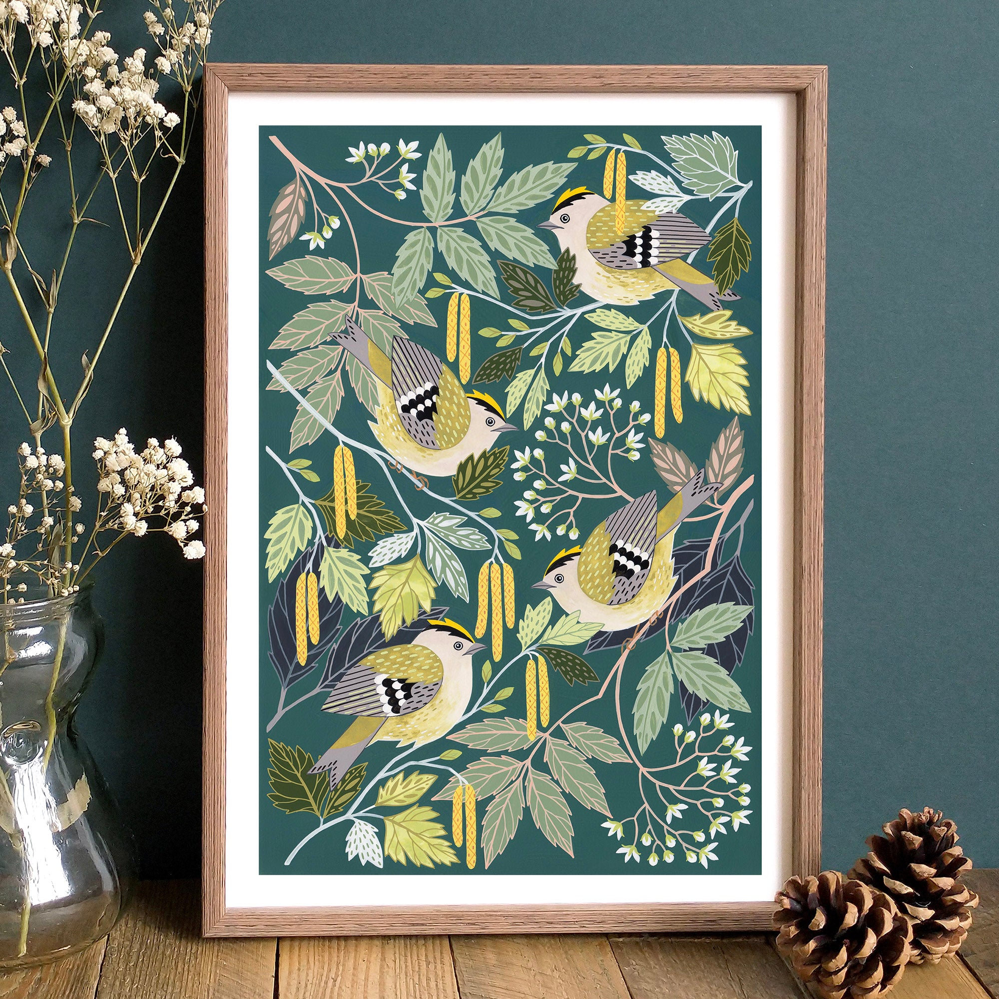 A framed art print featuring four goldcrests among green leaves and yellow catkins on a dark green background, styled on a wooden surface with dried flowers in a glass vase and pinecones against a dark teal wall.