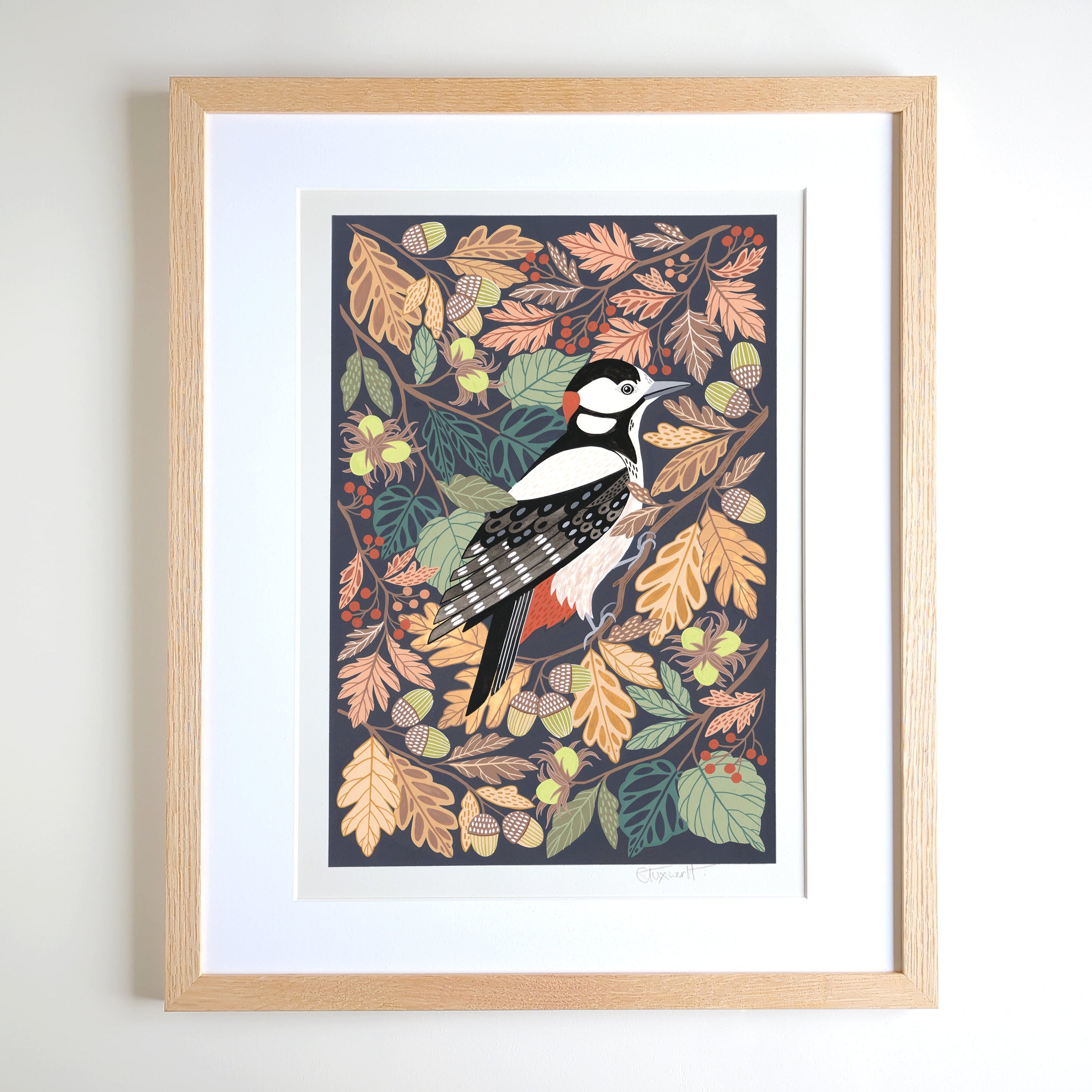 Framed art print featuring an illustration of a woodpecker surrounded by autumn leaves and acorns, displayed in a light oak frame with a white mount against a white wall.