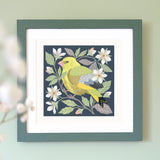 A framed painting featuring a greenfinch perched among white flowers and green leaves on a dark navy background. The painting is framed and displayed on a pale green wall.