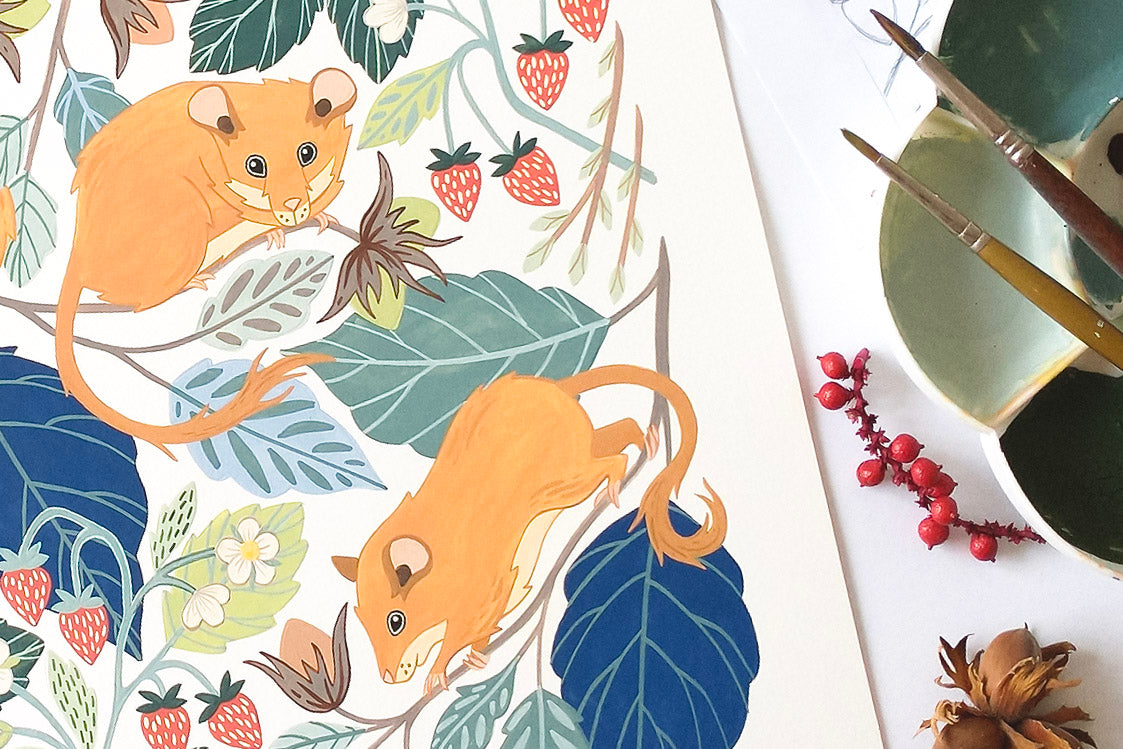 A flat-lay view of a painting of hazel dormice climbing among green leaves and red strawberries. To the right of the paper, a green ceramic paint palette with paintbrushes and small red berries on a white surface.