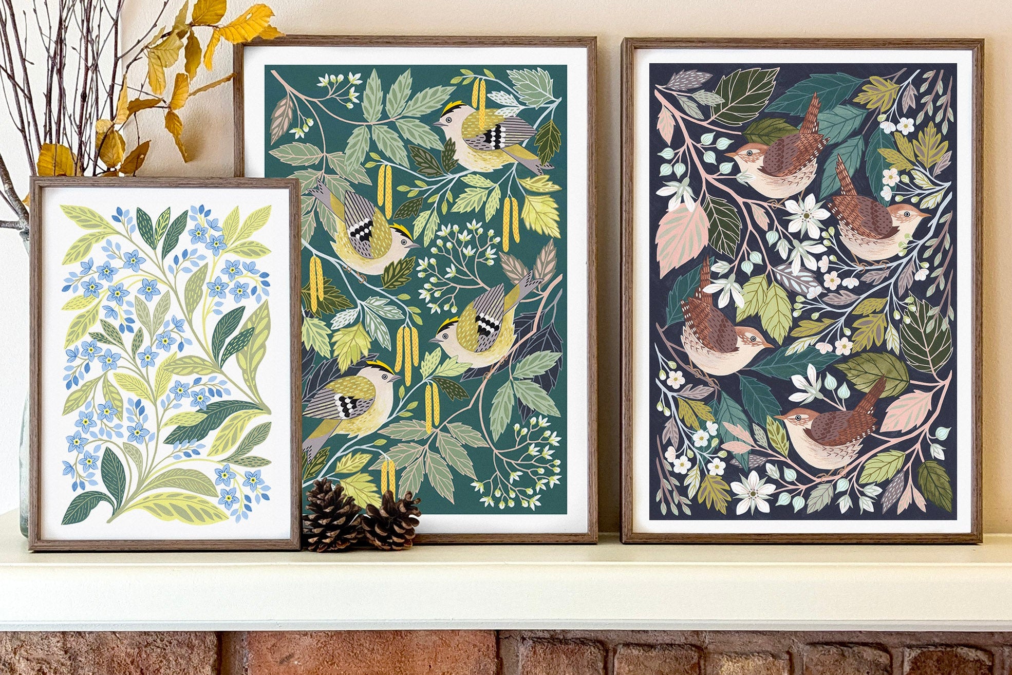 Three framed botanical and bird art prints displayed side-by-side on a white mantelpiece above a brick fireplace.