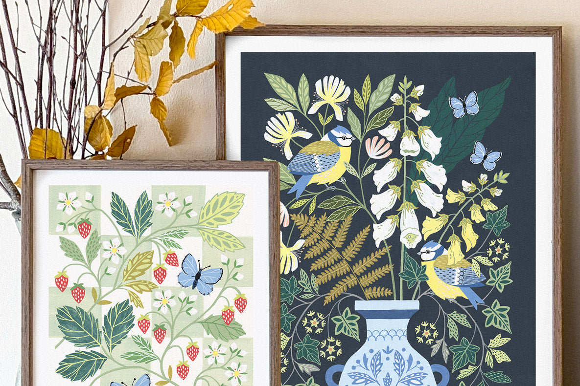 Two framed botanical art prints displayed side by side on a beige wall. The larger print on the right features birds, foxgloves, and green foliage on a dark navy background, while the smaller print on the left shows green leaves, red strawberries, and blue butterflies on a light green background.