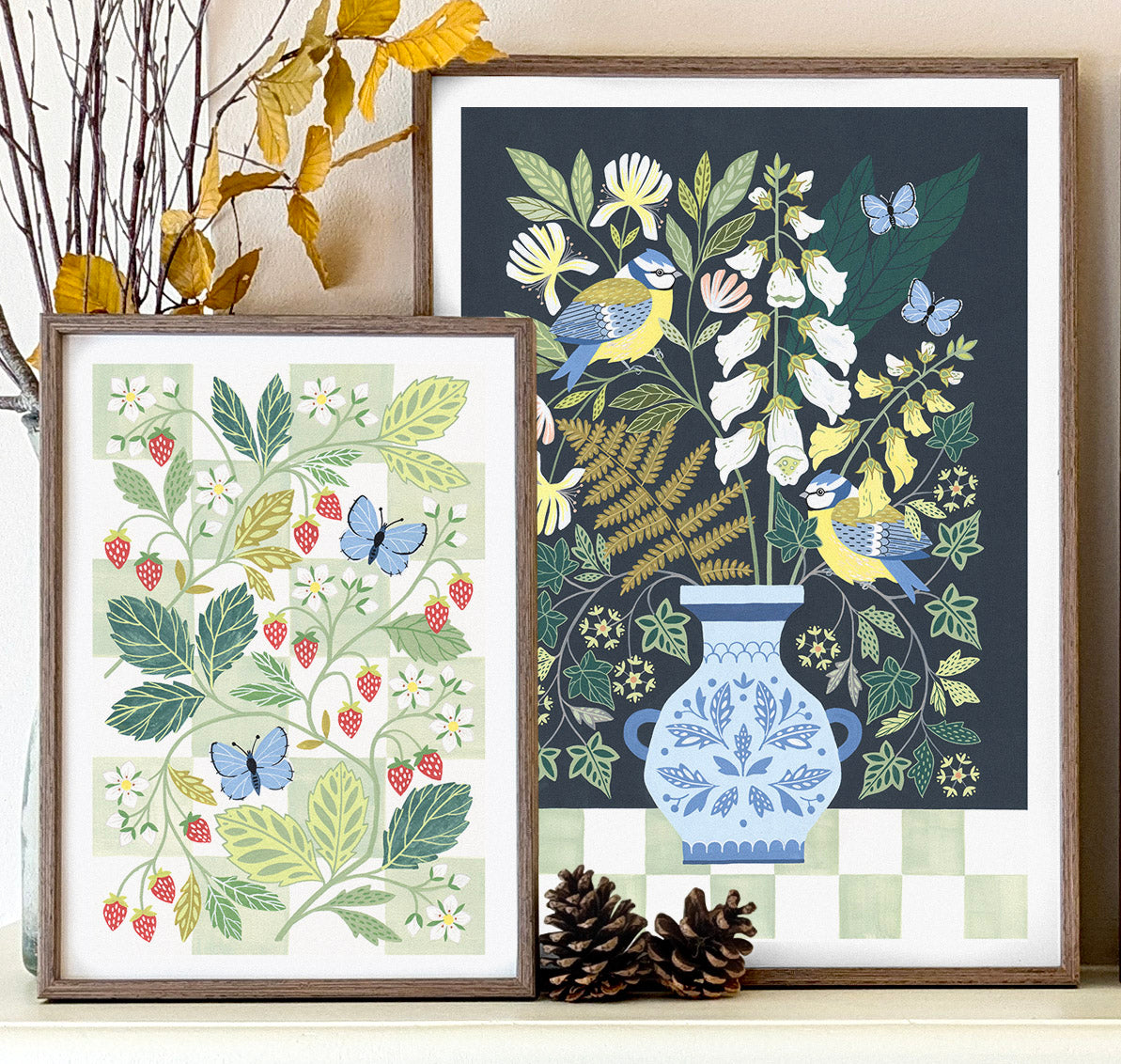 Two framed botanical art prints displayed on a shelf. The left print features strawberries and blue butterflies, and the right features birds and woodland foliage in a vase.