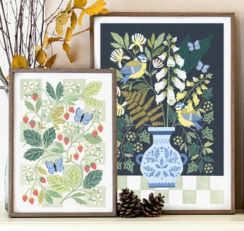 Two framed botanical art prints displayed on a shelf. The left print features strawberries and blue butterflies, and the right features birds and woodland foliage in a vase.